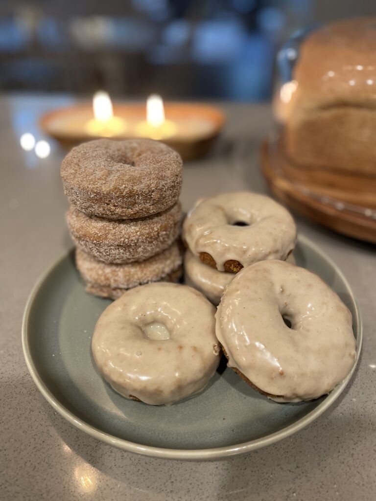 Fresh milled flour pumpkin donuts on a plate with a candle in the background, half coated in cinnamon sugar and half topped with maple cinnamon glaze.