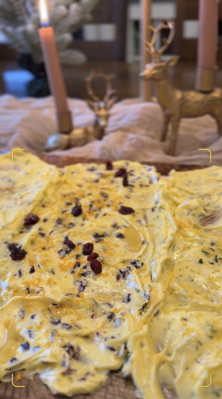A wooden butter board topped with three compound butters: roasted garlic and sage, cranberry orange with rosemary, and lemon thyme with flaky sea salt. Warm bread is torn and swiped through the butter on a softly lit holiday table.